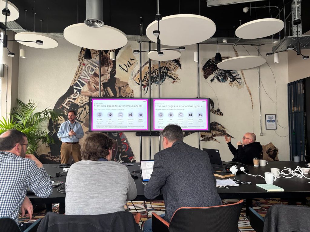 A big meeting room with circular shapes hanging from the ceiling, half a dozen men are sat at seats at the table, a man in a blue shirt and chinos is standing and talking in front of a screen, which says: From web pages to autonomous agents