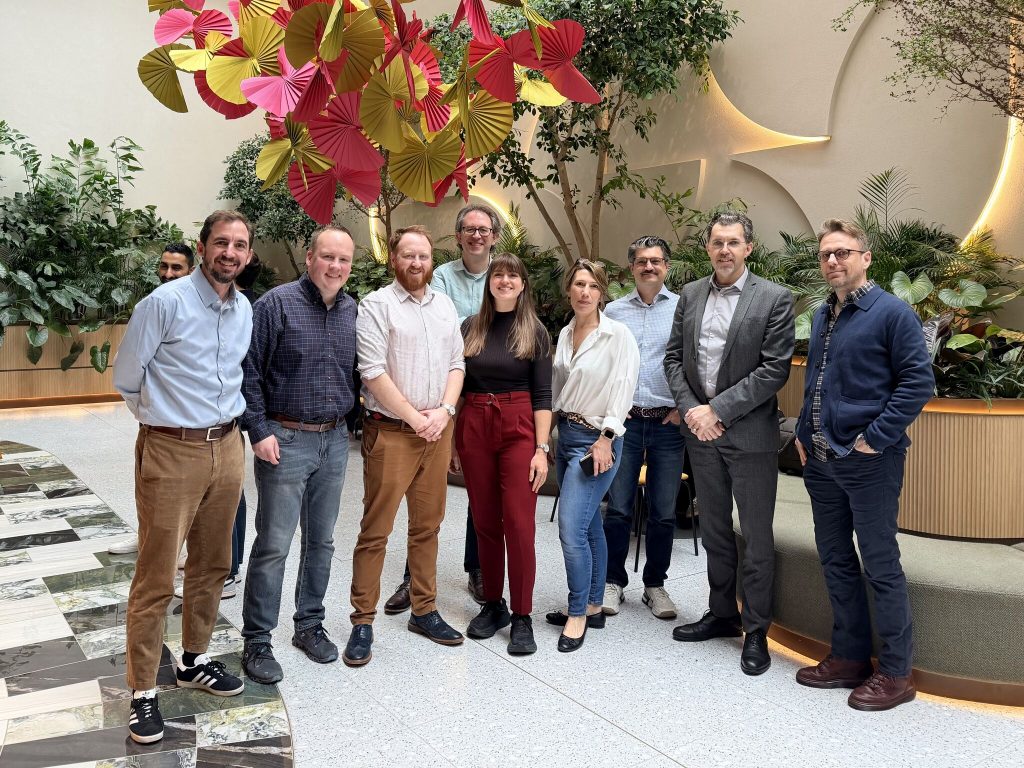 A group of 7 men and 2 women standing and smiling in a large open area of a smart office, with coloured paper fans above their heads.